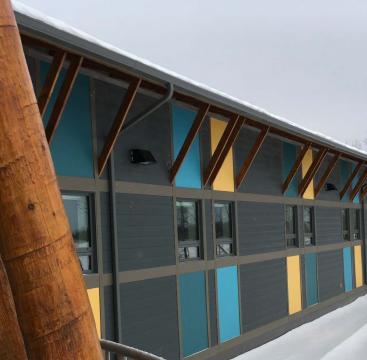 Kasabonika Lake First Nation, Image depicts the side of a black building with blue and yellow panels above and below the windows respectively. Season is winter and it's overcast.