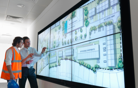 two men stand in front of a large display screen. on the screen is a map of a community with different buildings labelled. 