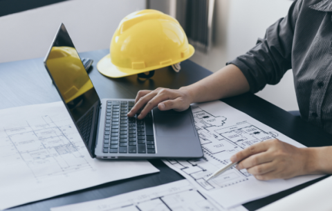 Close up of hands at a computer with a hard hat and blueprints nearby.